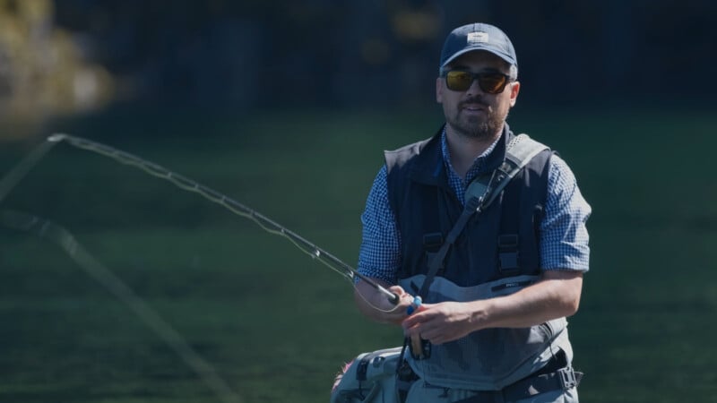 A man wearing sunglasses, a blue cap, and a fishing vest is fly fishing in a calm, green river, holding a fishing rod with both hands.