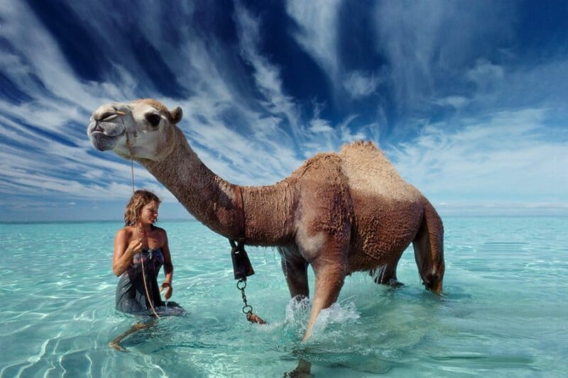A woman stands beside a camel in shallow, clear blue water under a dramatic sky with wispy clouds. The camel is on a lead and both appear relaxed in the scenic outdoor setting.