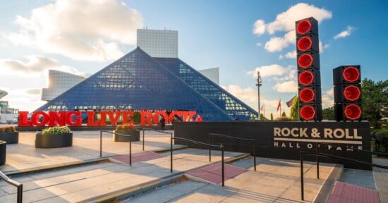 The Rock & Roll Hall of Fame in Cleveland, Ohio, features a glass pyramid structure, a large red "LONG LIVE ROCK" sign, and tall red speaker sculptures under a partly cloudy sky.