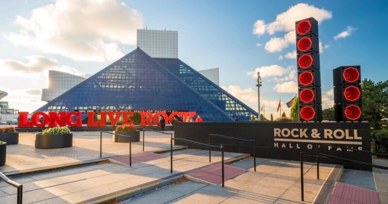 El Salón de la Fama del Rock and Roll en Cleveland, Ohio, presenta una estructura piramidal de vidrio, un gigante rojo "Larga vida al rock'n'roll" letrero y una alta escultura de altavoz rojo bajo un cielo parcialmente nublado.