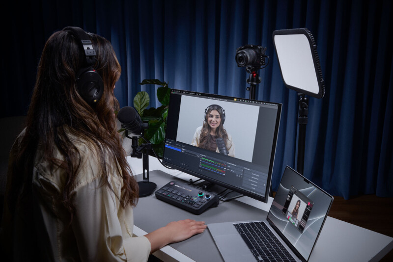 A woman wearing headphones sits at a desk with a microphone, streaming setup, and multiple screens, speaking to another woman shown on a monitor. Professional lighting and a camera are positioned in front of her.
