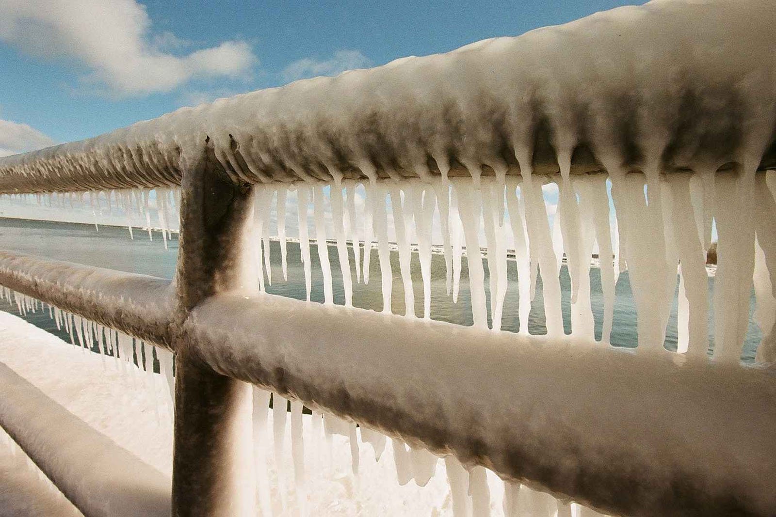 Icicles hang from a metal railing overlooking a body of water, formed by freezing weather. Snow and ice cover the ground, and the sky is partly cloudy with blue patches visible.