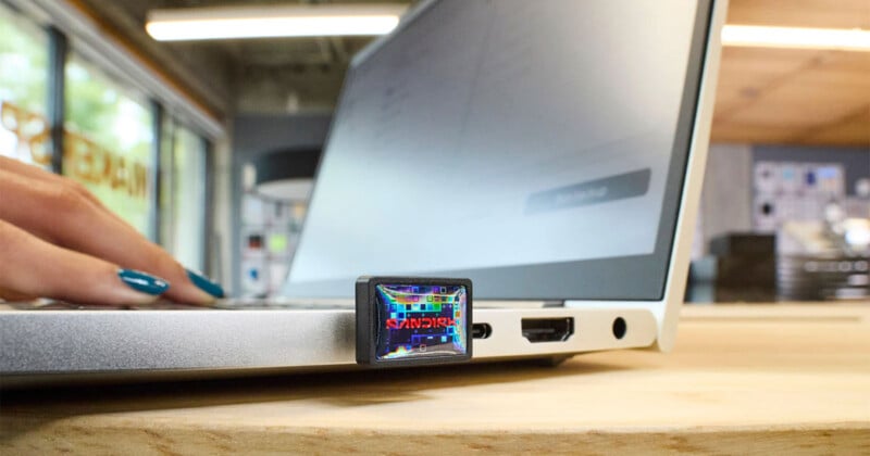 Primer plano de una mano usando una computadora portátil sobre una mesa de madera con una unidad flash USB conectada al puerto lateral de la computadora portátil. La unidad flash muestra una pantalla a color con texto y gráficos digitales.