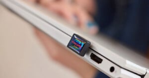 A close-up of a USB flash drive labeled "REGULARS" plugged into the side port of a silver laptop, with a blurred hand in the background.