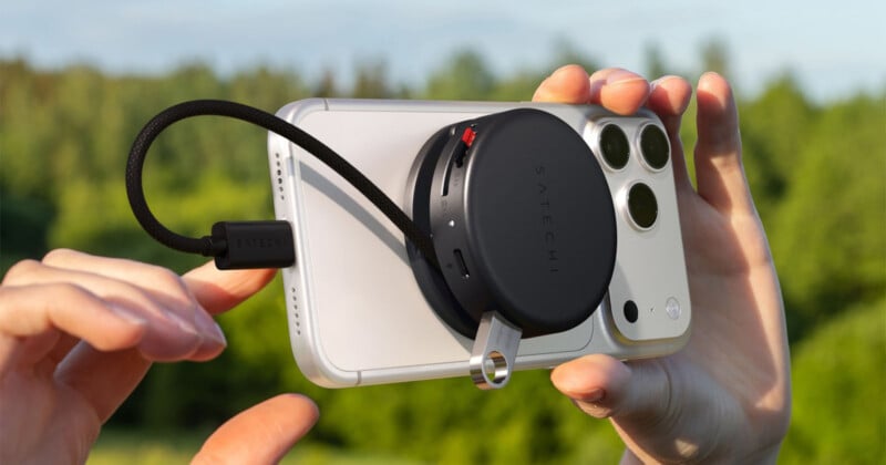 Un hombre sosteniendo un teléfono inteligente al aire libre con un dispositivo Satechi redondo conectado a la parte posterior conectado por un cable corto, contra árboles verdes borrosos en el fondo.