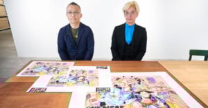 Two men sit at a table with colorful One Piece anime artwork spread out in front of them, against a plain white background. Both men are wearing glasses and formal jackets.