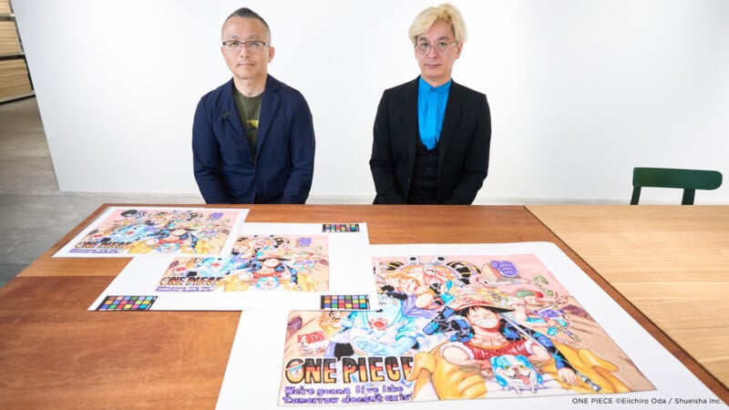 Dos personas sentadas en una mesa colorida "una pieza" Obras de arte y grabados. Estos grabados presentan personajes de anime con trajes brillantes. Los hombres miran a la cámara en una habitación moderna y luminosa.