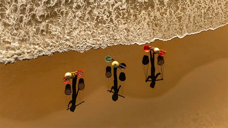 Vista aérea de tres coloridas sombrillas de playa que proyectan largas sombras sobre la arena húmeda cerca de la costa mientras las suaves olas se acercan desde la parte superior izquierda.