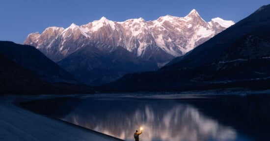 A person holding a light stands by a calm lake at dusk, with towering snow-capped mountains in the background reflecting on the water's surface.
