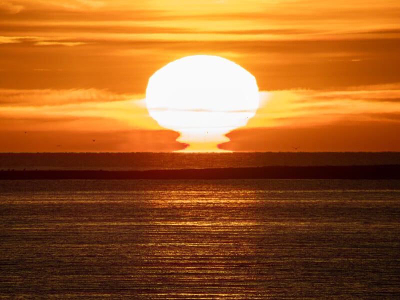El brillante sol anaranjado se eleva sobre el horizonte, proyectando reflejos dorados en las tranquilas aguas. Hilos de nubes cruzaban el cielo y una estrecha franja de tierra negra separaba el mar del cielo.