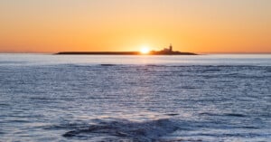 A lighthouse stands on a distant island as the sun rises behind it, casting an orange glow over a calm sea with gentle waves in the foreground.