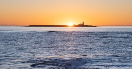 A lighthouse stands on a distant island as the sun rises behind it, casting an orange glow over a calm sea with gentle waves in the foreground.