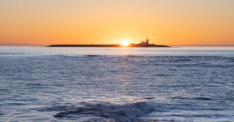 En una isla lejana hay un faro. El sol sale detrás del faro, proyectando un resplandor anaranjado sobre el mar en calma con suaves olas en primer plano.