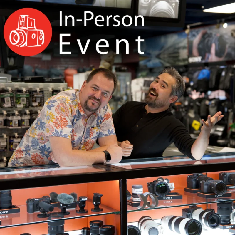 Two men stand behind a camera store counter, smiling and gesturing. Various cameras and lenses are displayed in glass cases. Text reads “In-Person Event” with a camera icon in the top left corner.