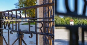A close-up of an iron gate with the inscription “Arbeit macht frei” and a view through the gate showing a watchtower and trees in the background at a historical site.