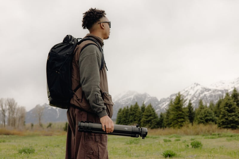 A person wearing sunglasses and a backpack stands outdoors on grass, holding a folded tripod, with snowy mountains and evergreen trees in the background under a cloudy sky.