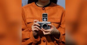 A person wearing an orange coat is holding a camera with both hands. The camera has a Ulanzi-branded flash or accessory mounted on top. The person's face is partially out of the frame.