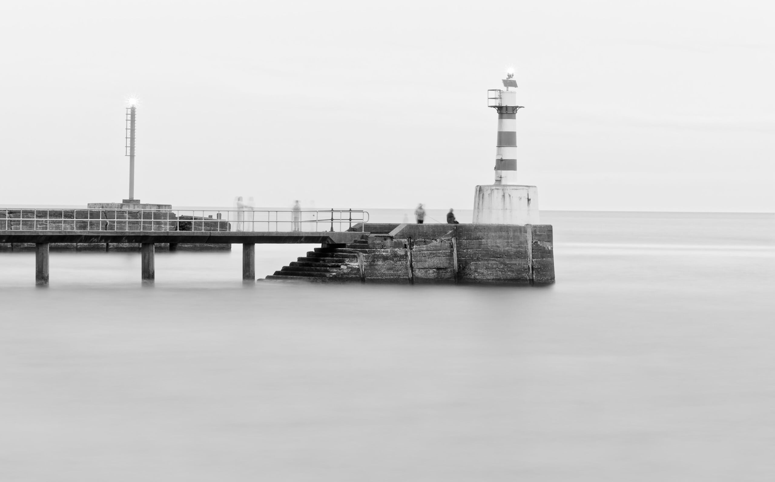 Una fotografía en blanco y negro que muestra un paisaje marino tranquilo con un muelle de piedra, un faro rayado, escaleras que conducen al agua y dos faros iluminados. La escena estaba tranquila y ligeramente brumosa.