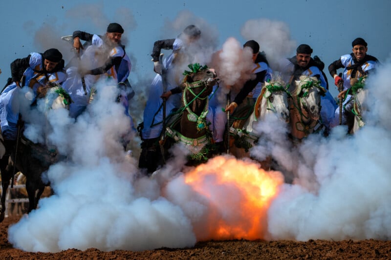 A group of men in traditional clothing ride decorated horses through thick smoke and fire during a cultural event or performance, with expressions of focus and excitement on their faces.