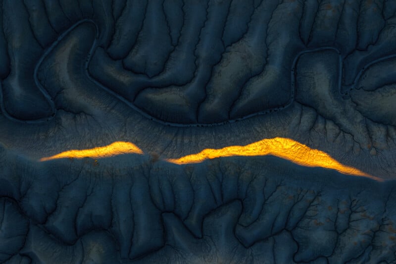 Aerial view of a rugged landscape with dark blue ridges and valleys, featuring a bright, jagged golden line of sunlight illuminating a central crevice.