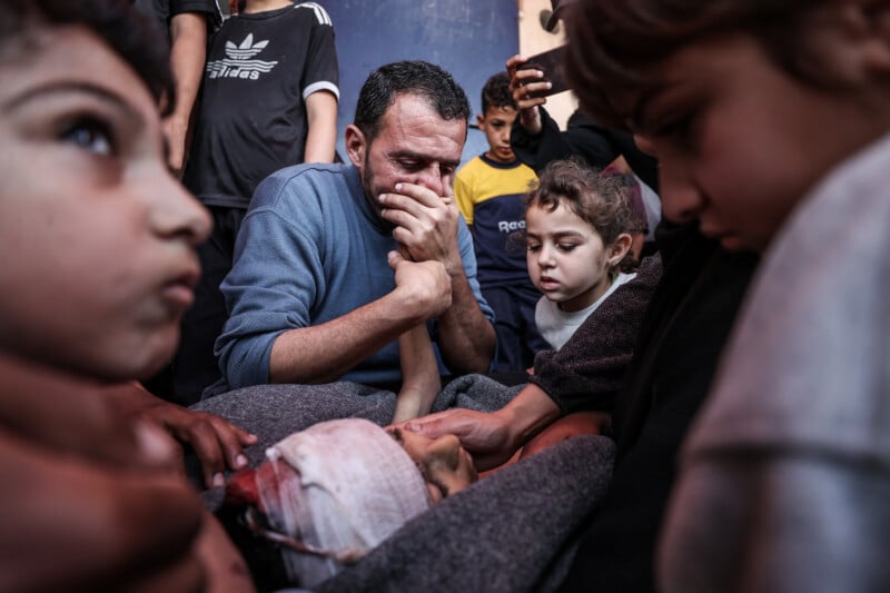 A grieving man covers his mouth in anguish, surrounded by children and others, as he mourns over a wrapped body with a bloodied bandage on the head, conveying intense sorrow and loss.