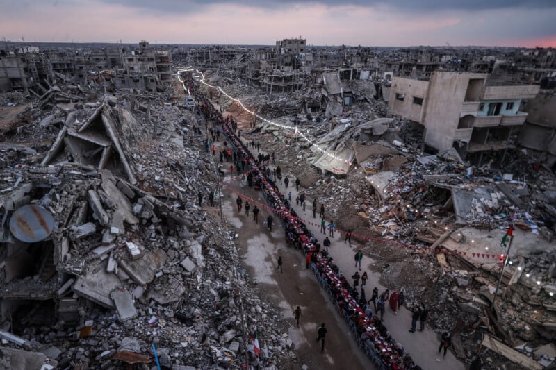 A long line of people walks through a street surrounded by heavily damaged and destroyed buildings in a city, with debris and rubble scattered everywhere and a cloudy sky above.