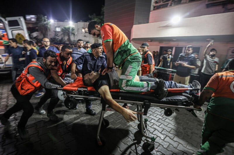 Emergency responders rush an injured person on a stretcher at night, surrounded by people in orange vests and uniforms, outside a building with bright lights. The scene appears urgent and chaotic.