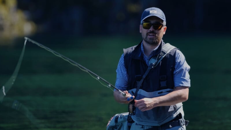 A man wearing sunglasses, a cap, and a fishing vest is fly fishing in a body of water with a greenish background. He is holding a fishing rod and appears focused on his activity.
