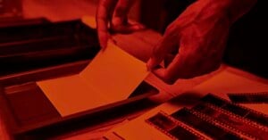 A person's hands develop a photo in a darkroom, holding photographic paper over a tray of liquid. Strips of film negatives and other photographic materials are visible on the wooden table under red lighting.