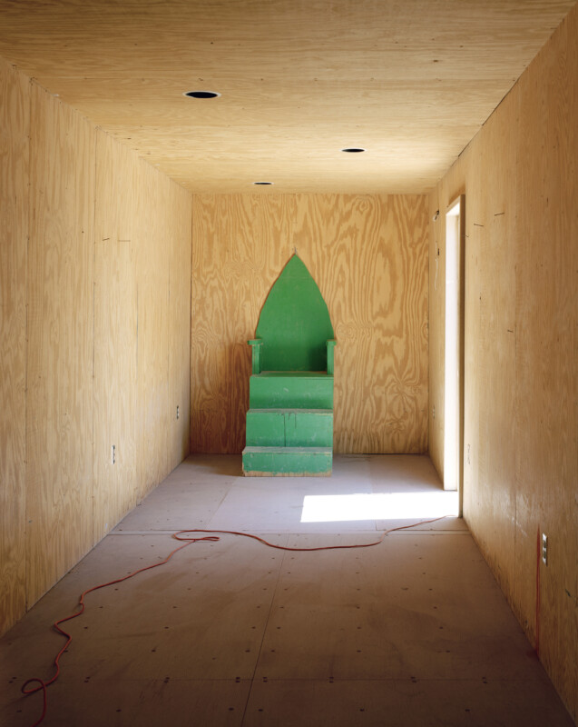 Una habitación pequeña y vacía con paredes y techo sin terminar, la luz del sol entrando por una puerta lateral. En la pared del fondo había una silla verde, parecida a un trono y con respaldo puntiagudo, con un cable de extensión naranja en el suelo.