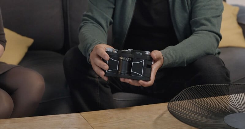 A person wearing a green jacket sits on a couch, holding a vintage black camera with both hands. The camera rests above a wooden table, and a modern wire bowl is visible in the foreground.