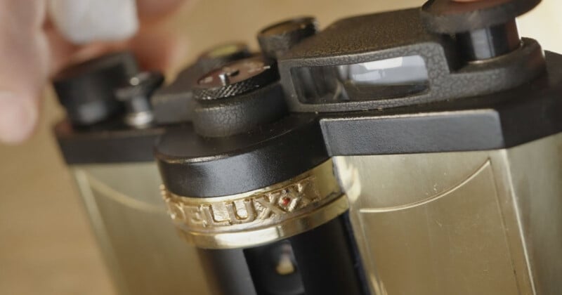 Close-up of a vintage twin-lens reflex camera with gold accents and textured black details, showing the viewfinder and part of the lens, as a hand adjusts a control knob.