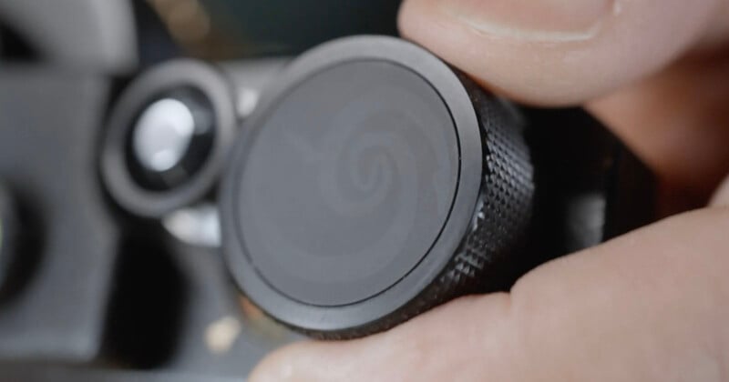 A close-up of a person’s fingers adjusting a textured black control dial on a camera or electronic device. The dial has a subtle spiral pattern on its surface.