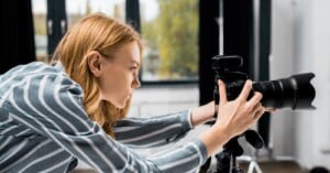 A woman with red hair and a striped shirt adjusts a professional camera on a tripod indoors, focusing intently on her shot. Large windows with blurred greenery are visible in the background.
