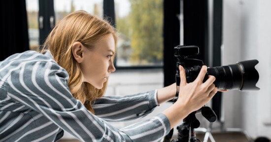 A woman with red hair and a striped shirt adjusts a professional camera on a tripod indoors, focusing intently on her shot. Large windows with blurred greenery are visible in the background.