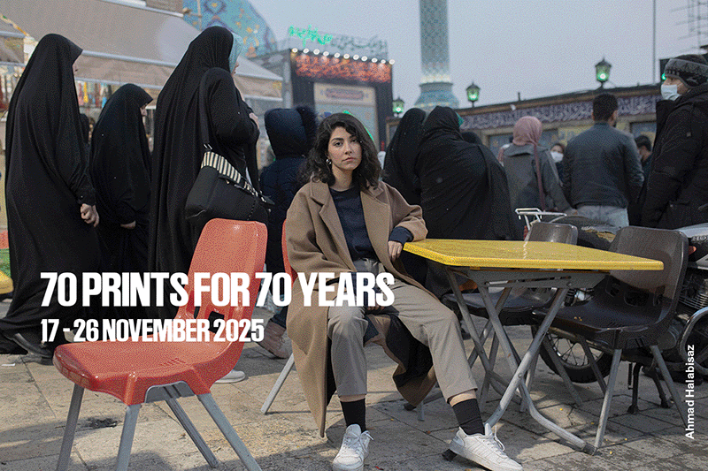 Una mujer vestida de color canela está sentada en una silla roja en un zoco, rodeada de sillas vacías y gente vestida de negro. El texto dice, "70 años impresos, del 17 al 26 de noviembre de 2025." Crédito de la foto: Ahmed Halabisaz.