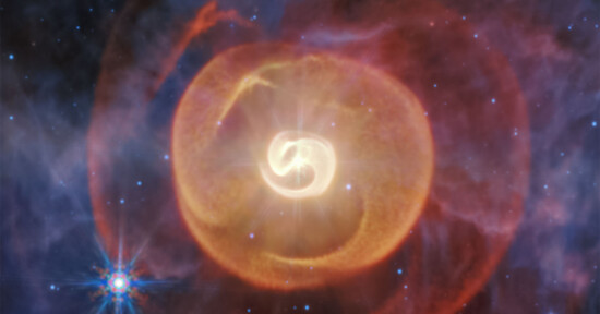 A bright, glowing star surrounded by swirling red, orange, and yellow gas clouds in space, with distant stars scattered across a dark background and a blue star in the lower left corner.