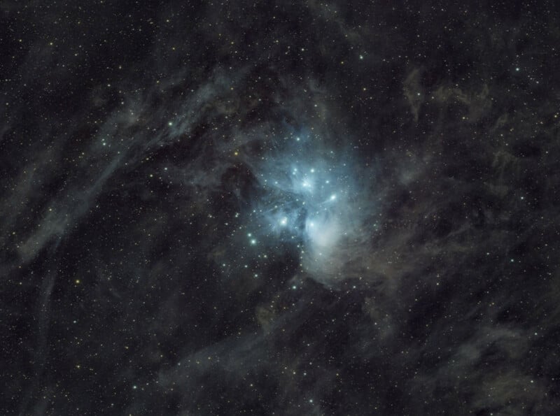 Nebulosas de color azul brillante brillan en un campo estelar oscuro en el espacio, rodeadas por un tenue polvo interestelar e innumerables estrellas pequeñas esparcidas por la imagen.