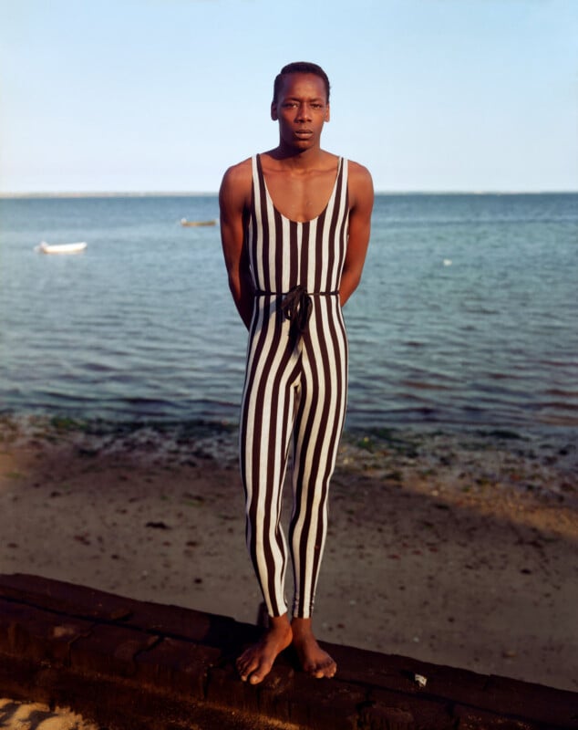 A person stands barefoot on a beach in front of the ocean, wearing a black and white vertically striped sleeveless jumpsuit. Their arms are behind their back, and the sky is clear.