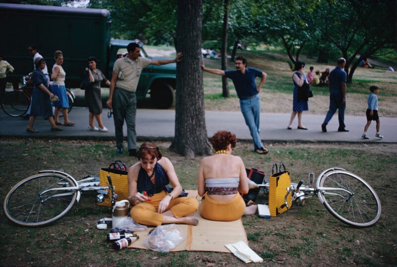Two women sit on a mat next to their bicycles, having a picnic in a park. People walk and stand nearby, and trees and grass surround the scene on a sunny day.