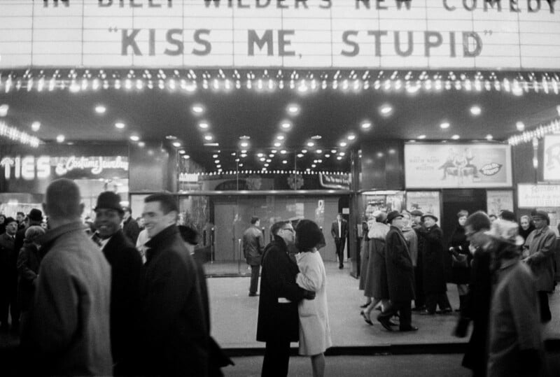 A black-and-white photo of a busy street outside a theater showing "Kiss Me, Stupid." People walk by, while a couple stands in the center, kissing under the theater marquee’s lights.