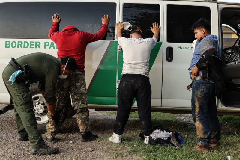 A Border Patrol officer searches three men with their hands against a white van labeled "Border Patrol." The men appear muddy and are being detained; clothing and bags are on the ground nearby.