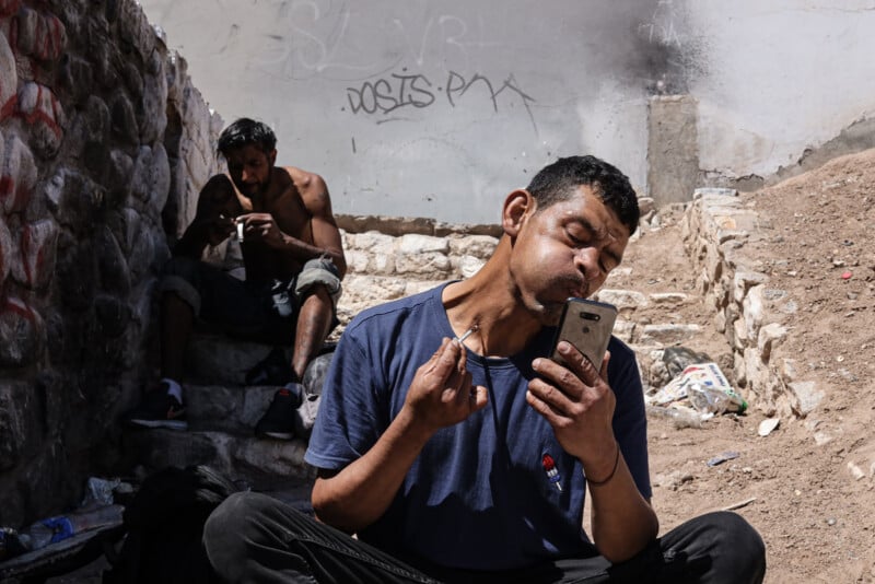 A man sits on the ground holding a phone and injecting his neck, while another man sits on stone steps in the background preparing to inject himself. The area is rundown, with graffiti on the wall and scattered trash.
