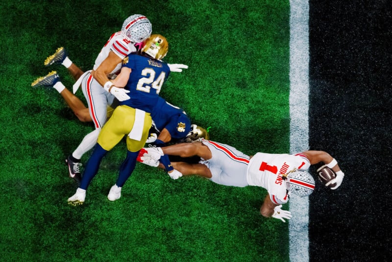 A football player in a white uniform reaches across the goal line for a touchdown while defenders in blue and gold uniforms attempt to stop him. The action takes place on a green field with a black and white end zone.