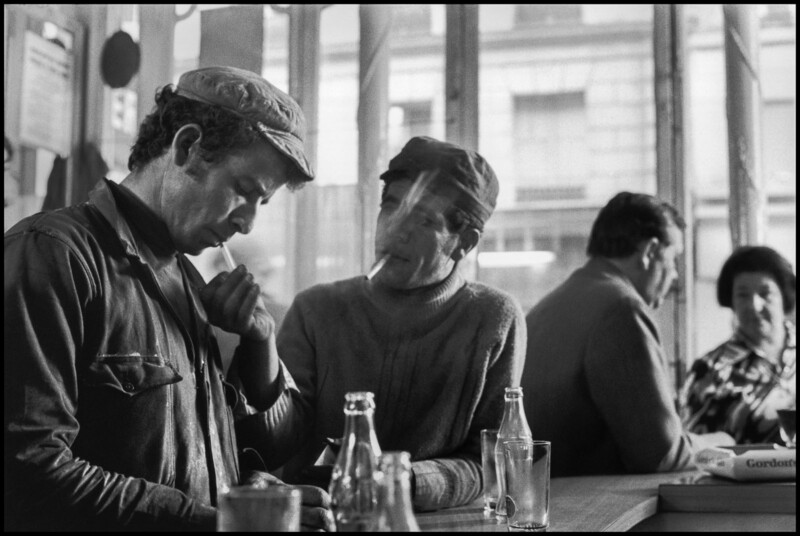 Two men sit at a café counter; one lights the other's cigarette. Both wear work clothes and caps. Empty glass bottles and cups are on the counter. In the background, two other patrons sit and talk near the window.