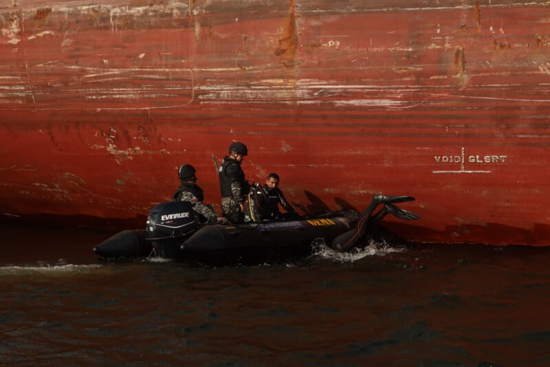 Three uniformed individuals in a black inflatable boat patrol beside the rusty hull of a large ship on the water. The boat is labeled "OMA" and equipped with an outboard motor.