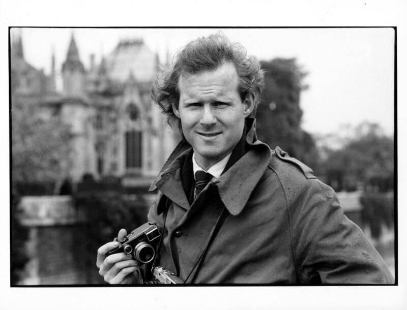 A man wearing a trench coat and tie holds a camera, standing outdoors in front of a historic building with pointed arches and towers. The photo is in black and white.