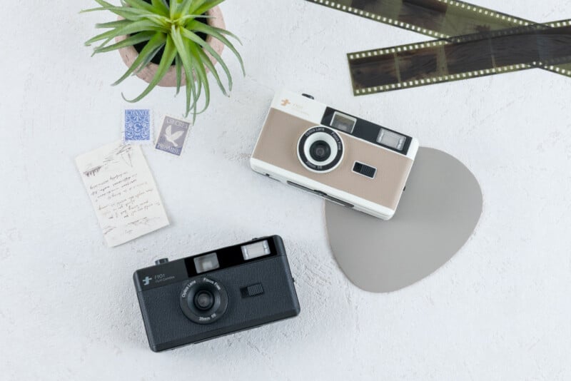Dos cámaras antiguas, una negra y otra blanca, se encuentran sobre una superficie clara, junto a una pequeña planta verde en una maceta, una tira de película, una nota escrita a mano y dos sellos.