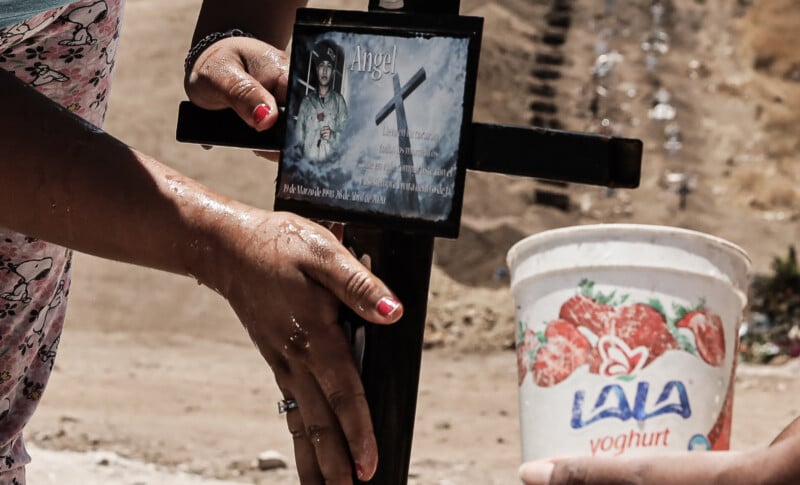 A person touches a black cross with a memorial photo and message, while another hand holds a white Lala yogurt cup. The background is a dry, sandy area with blurred steps or graves.