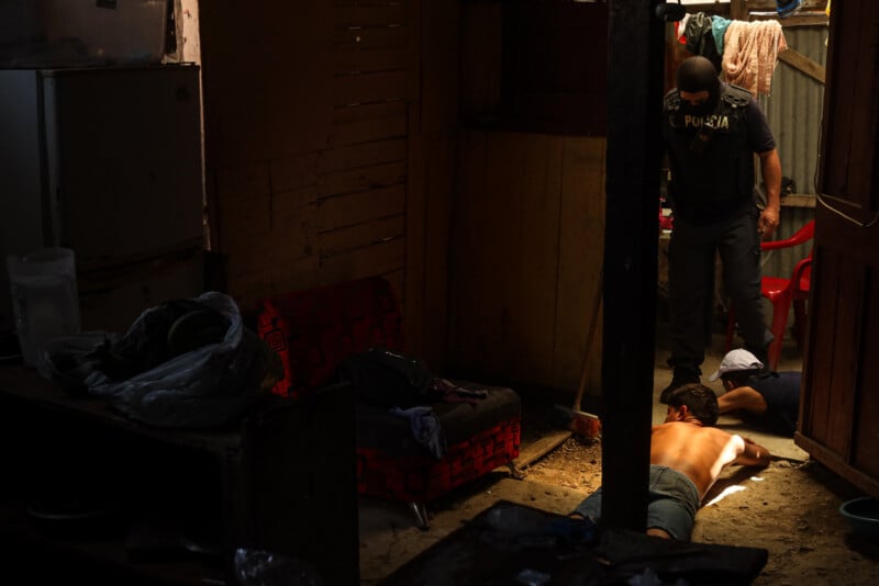 A police officer stands in a dimly lit, cluttered room while two men lie face down on the floor. One man is shirtless. Sunlight streams through an open door, illuminating part of the scene.